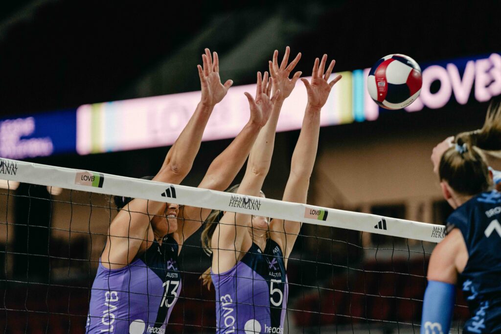 LOVB pro volleyball players jump above the net to spike a ball.