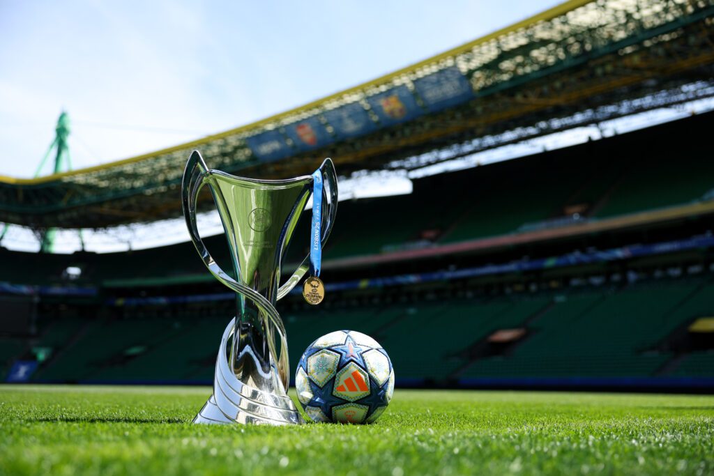 The 2024/25 UEFA Champions League trophy, medal, and game ball sit on the Estádio José Alvalade pitch in Portugal.