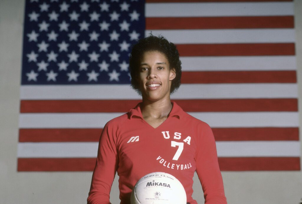 1984 Olympian Flo Hyman poses in front of a US flag holding a volleyball.