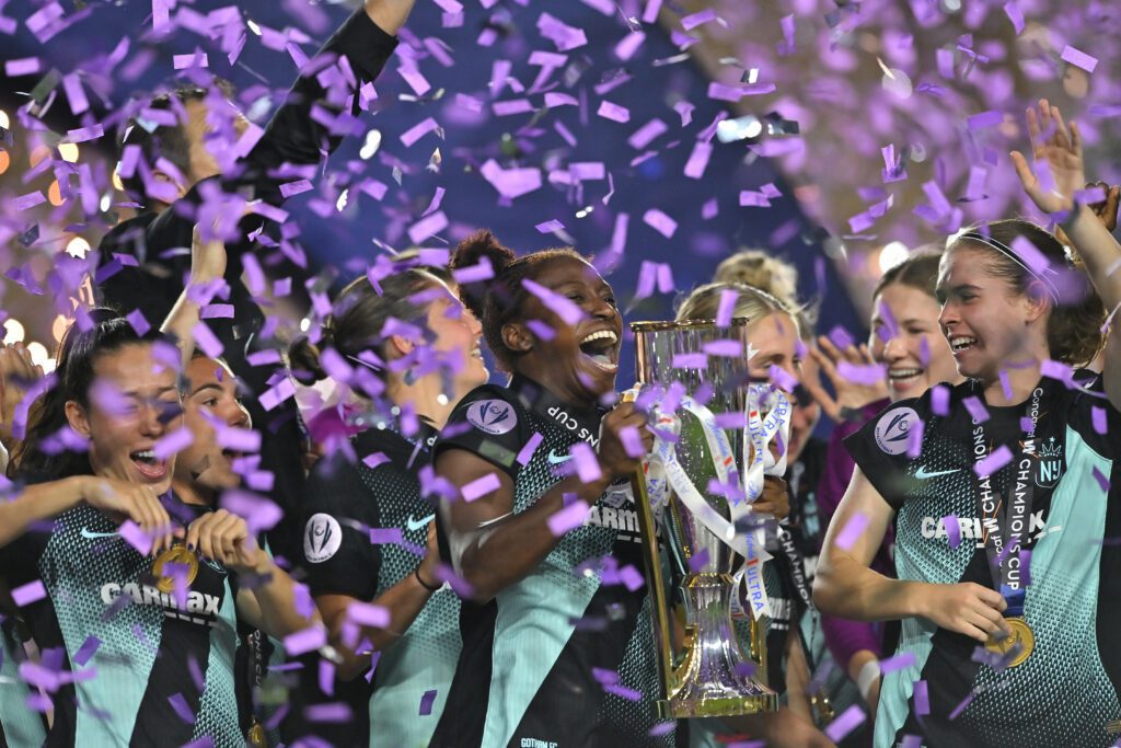 Gotham defender Mandy Freeman lifts the 2024/25 Concacaf Champions Cup and celebrates with her teammates as purple confetti falls.